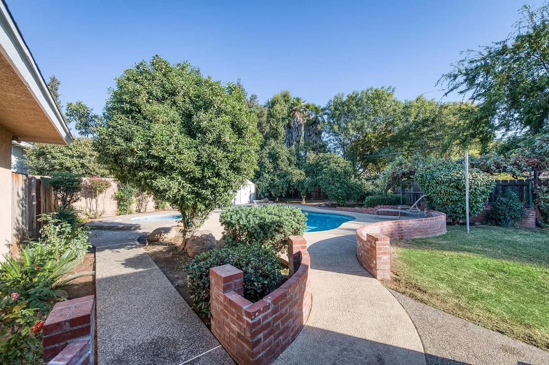 The photo shows a landscaped backyard with a swimming pool, curved concrete walkways, brick retaining walls, shrubs, and mature trees.