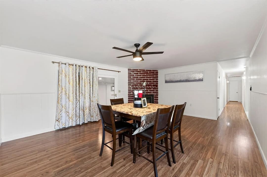 A long, unfurnished-looking dining or living area with wood-look flooring, white walls, and a brick fireplace at the far end.