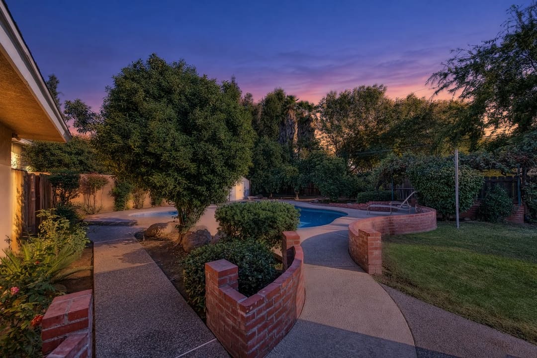 The photo shows a landscaped backyard with a swimming pool, curved concrete walkways, brick retaining walls, shrubs, and mature trees.