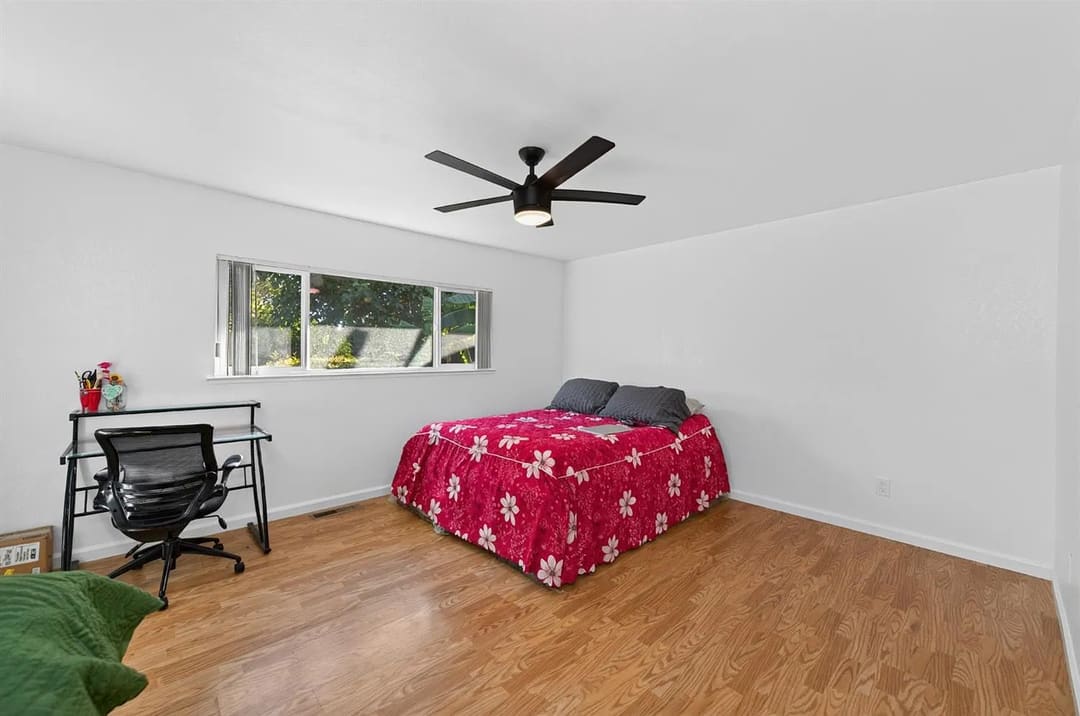 A simple bedroom with white walls, wood-look flooring, a bed with a bright pink floral bedspread, and a small desk with an office chair.