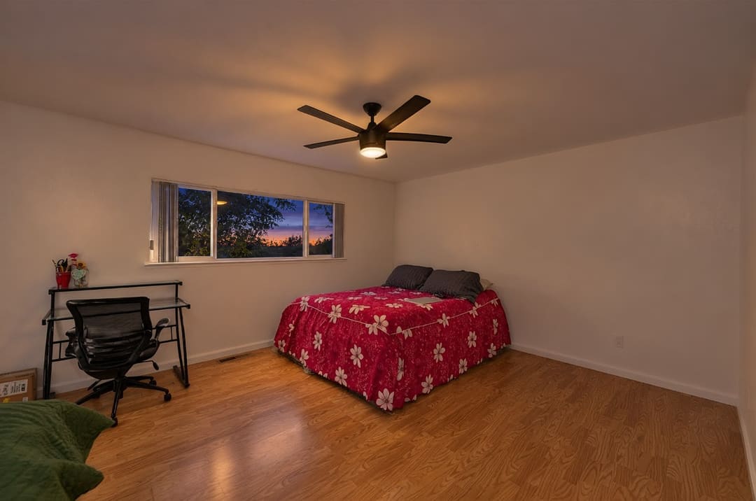 A simple bedroom with white walls, wood-look flooring, a bed with a bright pink floral bedspread, and a small desk with an office chair.