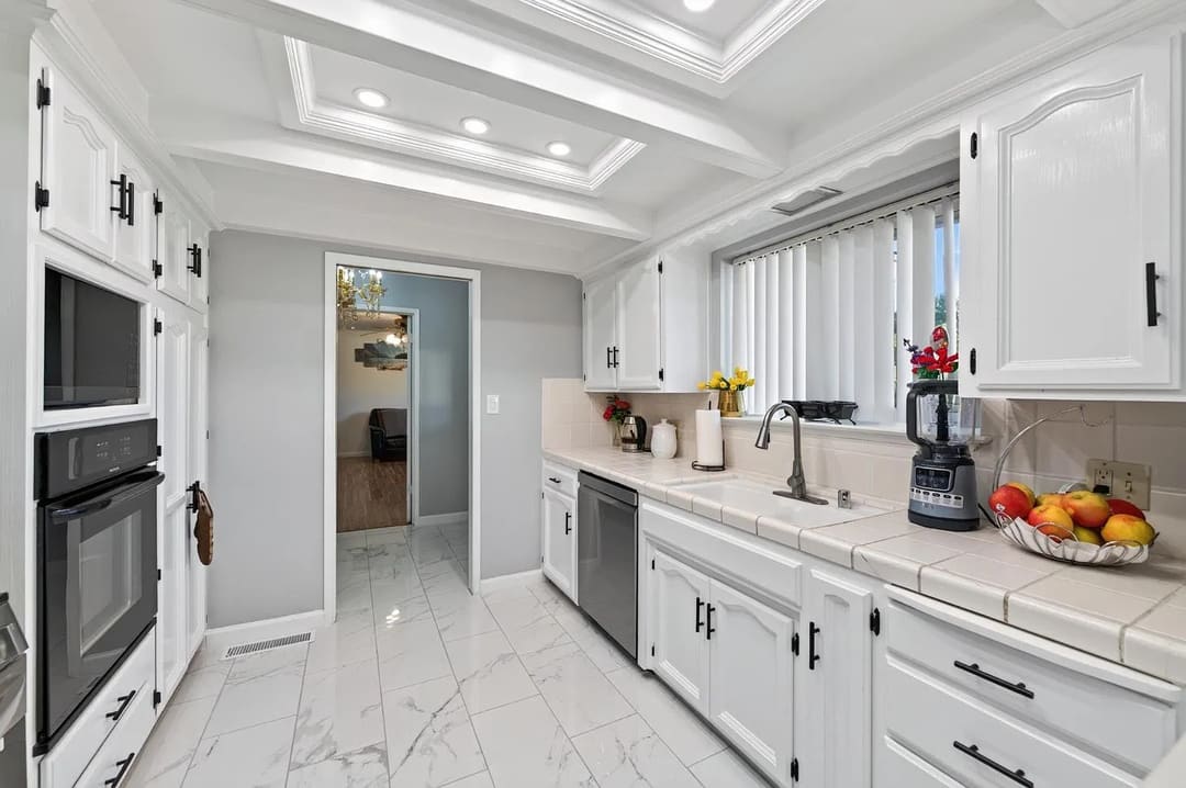 A bright narrow kitchen with white cabinets on both sides, a tiled floor, and a doorway at the far end leading to another room.