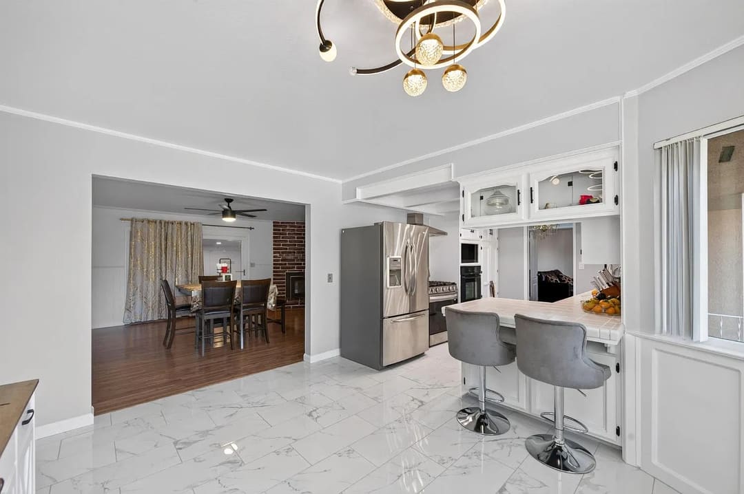 A bright kitchen with glossy white marble-look tile flooring, white walls, and a breakfast bar with two gray stools.